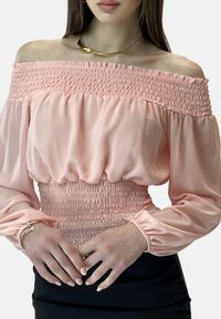 Blouse rose à épaules dénudées avec smocks, manches longues bouffantes. Tissu doux et léger avec un corps texturé et une taille élastiquée.