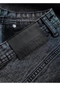 Les jeans en denim affichent une couleur gris foncé avec une surface texturée. Une patch en cuir noir embossé avec "OMBRE" est fixé à la taille.