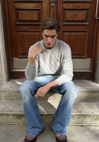 Mladý muž v sivom svetri a modrých džínsoch sedí na betónových schodoch pred drevenými dverami a pozerá sa dole s premýšľavým výrazom.