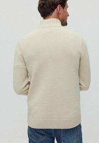 Beige stickad tröja med hög krage, ribbade mudd och nederkant. Bakifrån visar den en jämn yta och en figurnära silhuett.