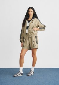 Smėlinio atspalvio sportinė bėgimo striukė ir šortai su juodomis juostelėmis, derinami su balta marškinėliais ir sidabrinėmis sportinėmis avalynėmis. Lygus audinys, laisvas siluetas.