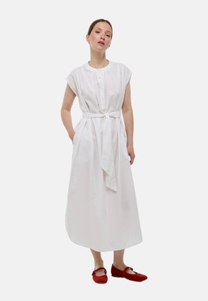 Robe en coton blanche à manches courtes, avec un devant à boutons, une taille cintrée par une ceinture et des poches latérales. Associée à des chaussures plates rouges.