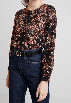 Femme portant un haut noir à manches longues avec un motif de branches florales orange, rentré dans un jean bleu foncé avec une ceinture noire.
