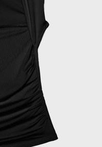 Section de tissu noir avec fronces rassemblées le long d'une couture verticale sur un fond gris clair.