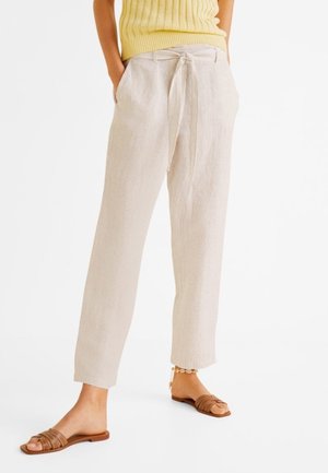 Frau trägt beige gerippte Hose mit gebundener Taille, ein gelbes ärmelloses Oberteil, braune flache Sandalen und ein Fußkettchen vor weißem Hintergrund.