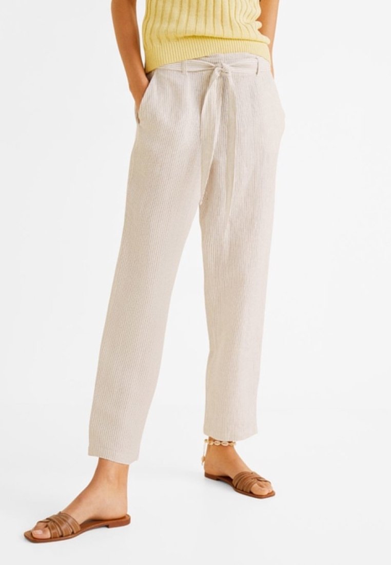 Femme portant un pantalon beige côtelé avec une taille nouée, un haut sans manches jaune, des sandales plates marron et un bracelet de cheville sur fond blanc.