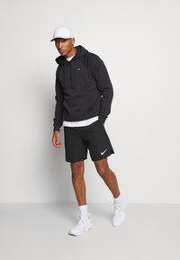 Sudadera con capucha negra y bolsillo frontal, combinada con pantalones cortos negros que presentan un logo blanco de Nike. Zapatillas deportivas blancas y una gorra blanca.