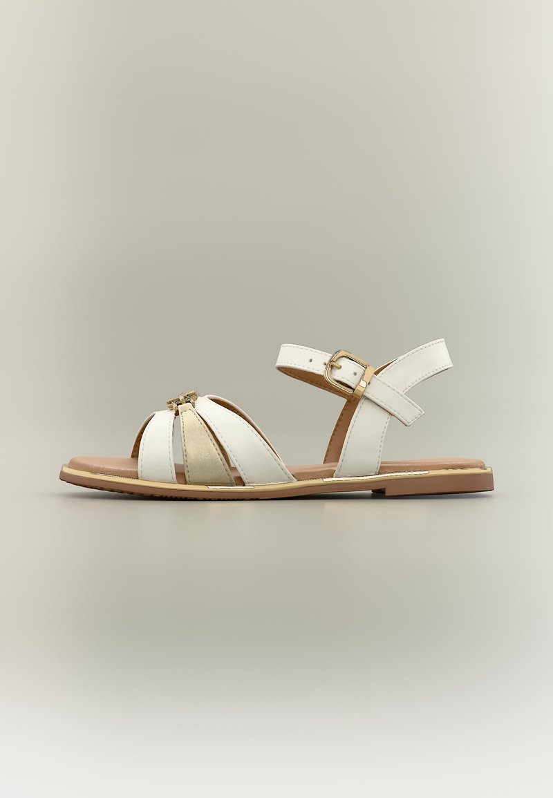 Λευκό επίπεδο σανδάλι με ανοιχτό δάχτυλο, λουρί στον αστράγαλο με χρυσή πόρπη και διακοσμητική χρυσή λεπτομέρεια στα μπροστινά λουριά, σε ουδέτερο φόντο.