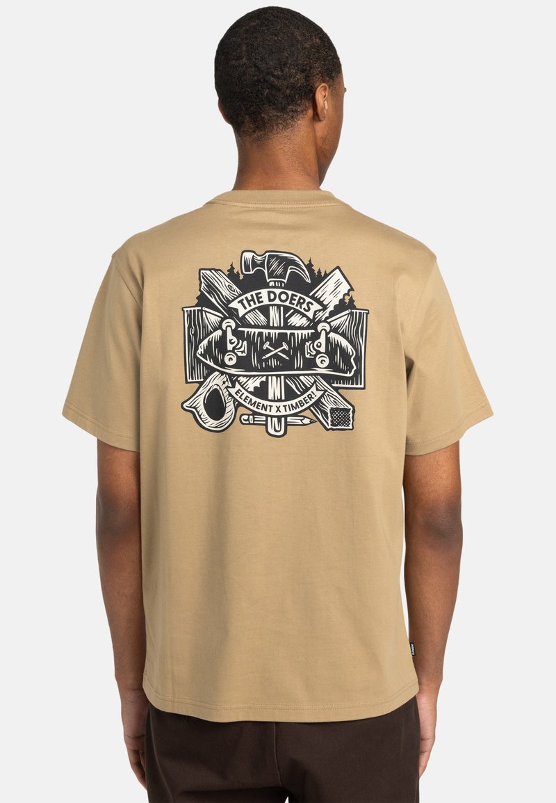Beige bomulds t-shirt med et stort sort grafisk design på ryggen, inklusive værktøj og teksten "DE GØRENDE". Korte ærmer, afslappet pasform.
