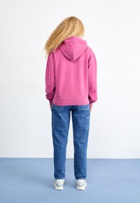 Felpa in pile di un acceso colore rosa con cappuccio, abbinata a pantaloni cargo in denim blu con tasche laterali, su sfondo neutro.