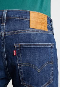Primer plano de un bolsillo trasero de unos jeans Levi's azul con costura característica, etiqueta roja de Levi's y parche de cuero marrón en la cinturilla.