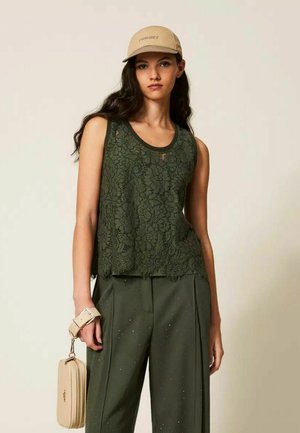 Donna con cappellino beige, top senza maniche in pizzo verde scuro, pantaloni abbinati con piccoli fori e che tiene una borsa beige per la tracolla.