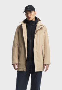 Beige buitenjack met een capuchon, voorzien van een zwart ritsjack aan de binnenkant. De stof lijkt zacht en waterafstotend te zijn.