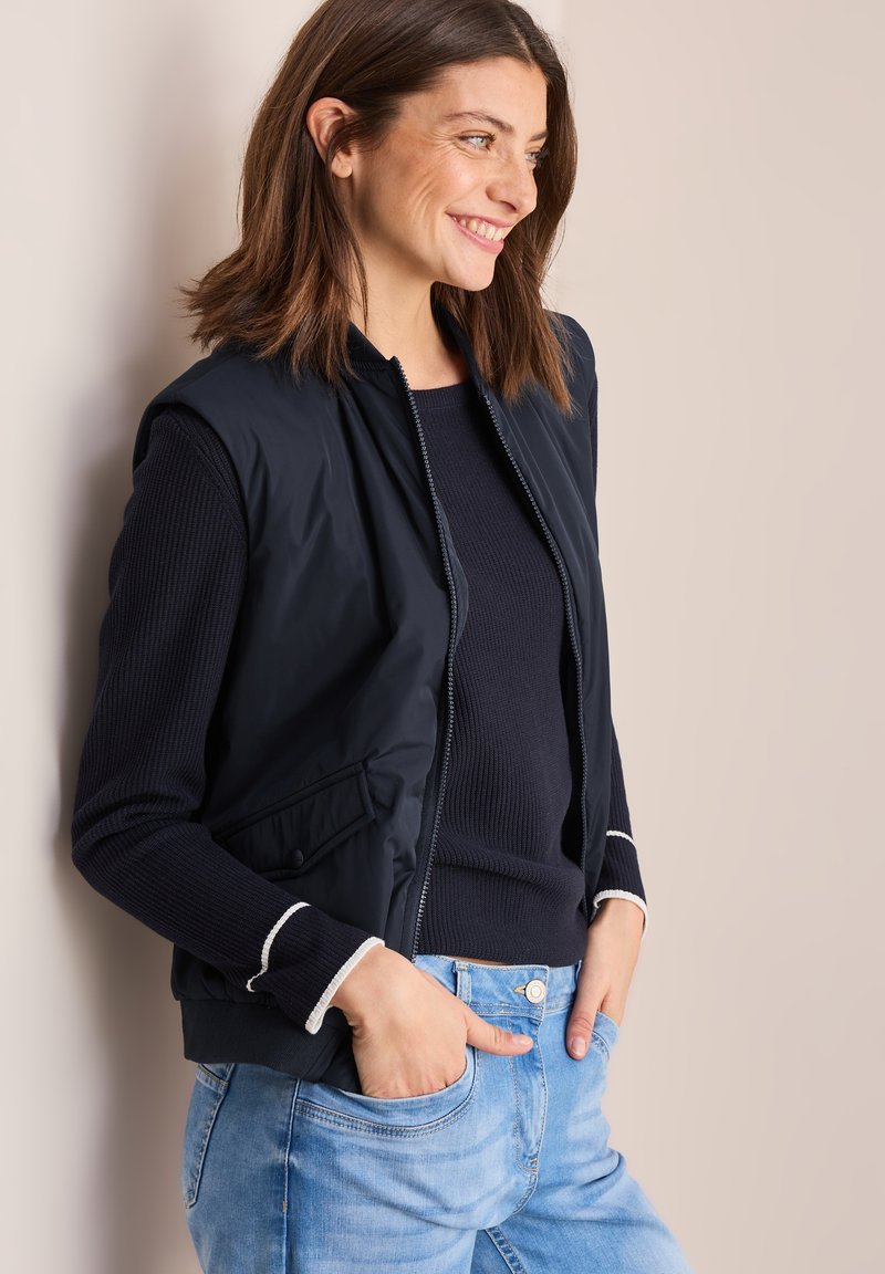 Gilet navy avec fermeture éclair, poches latérales et bord blanc aux poignets, porté sur un haut en maille foncé, associé à un jean bleu clair.