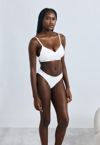 Biely set spodnej bielizne pozostávajúci z braletky a nohavičiek v štýle bikín. Materiál je hladký s priliehavým strihom. Pás s logom pridáva detail.