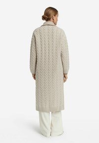 Lång, beige kabelstickad cardigan med krage, ribbade ärmslut och nederkant. Texturen har ett upphöjt mönster som ger ett mysigt utseende.