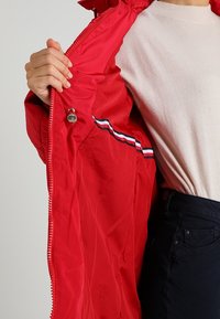 Veste matelassée rouge avec extérieur lisse, fermeture éclair et doublure rayée bicolore. Comprend un bouton-pression et un intérieur aux détails minimalistes.