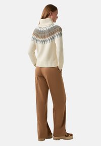 Vrouw in een crèmekleurige coltrui met een grijs en bruin gestreepte yoke, bruin wijde broek en bruine wandelschoenen, staand in zijaanzicht.