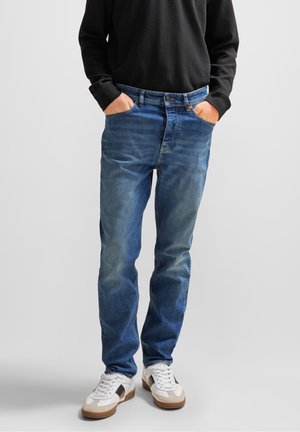 Person trägt blaue Jeans, weiße Sneaker und ein schwarzes Langarmshirt, steht mit den Händen in den Taschen vor einem schlichten Hintergrund.
