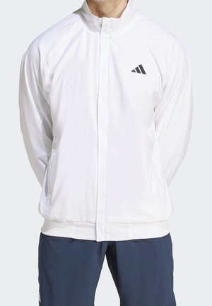 Man draagt een wit jack met ritssluiting en een zwart Adidas-logo op de borst, en donkerblauwe broek, staand met handen achter de rug.