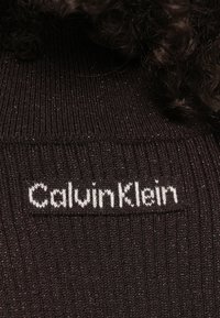 Hnedý pletený top s rebrovanou textúrou a bielym logom "CalvinKlein". Tkanina obsahuje jemné trblietavé nite po celej ploche.