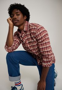 Camicia a quadri rossa e bianca con maniche lunghe, indossata con pantaloni di denim blu arrotolati. Sono visibili anche scarpe da ginnastica blu e calzini a fantasia.