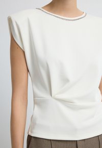 Biała dopasowana bluzka z delikatnym połyskiem, strukturalnymi ramionami, marszczeniem w pasie oraz zdobionym dekoltem z małymi kryształkami.