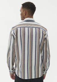 Moški, oblečen v srajco s dolgimi rokavi in gumbi, z navpičnimi črtami v beli, mornarsko modri, peščeni in olivni barvi, obrnjena stran od bele ozadje.