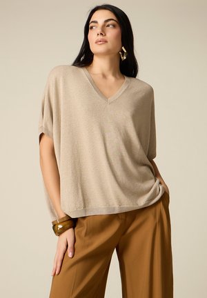 Donna con top beige largo a scollo a V e maniche corte, pantaloni marroni a gamba larga, orecchini geometrici dorati e bracciale marrone.
