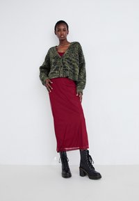 Zelený a černý pletený cardigan přes červené šaty s krátkým střihem. Černé šněrovací boty s masivním podpatkem dokončují outfit.