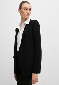 Czarny żakiet o dopasowanym kroju, z pojedynczym zapięciem na guzik i bocznymi kieszeniami; w zestawieniu z białą koszulą z kołnierzykiem. Gładka tekstura materiału.