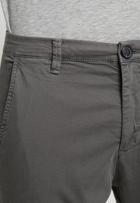Gros plan sur la taille d'un pantalon gris avec bouton, passant de ceinture et poche latérale, porté avec une chemise gris clair.