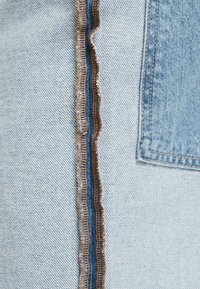 Ljusblå denimjeans med kontrasterande brun söm längs sidsömmen, med en strukturerad yta och detaljerade lapplommer.