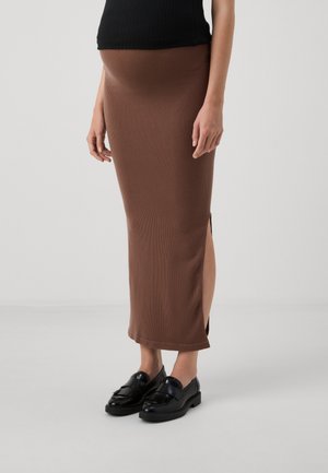 Smeđa rebrasta maxi suknja s bočnim prorezima, uparena s crnim loafericama. Outfit sadrži oblikovanu pojasnicu za čvrsto pristajanje.