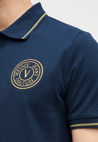 Κοντινό πλάνο ενός μπλε πόλο πουκάμισου με χρυσή διακόσμηση στη γλώσσα και το μανίκι, με ένα χρυσό κεντητό έμβλημα λογότυπου Versace Jeans Couture.