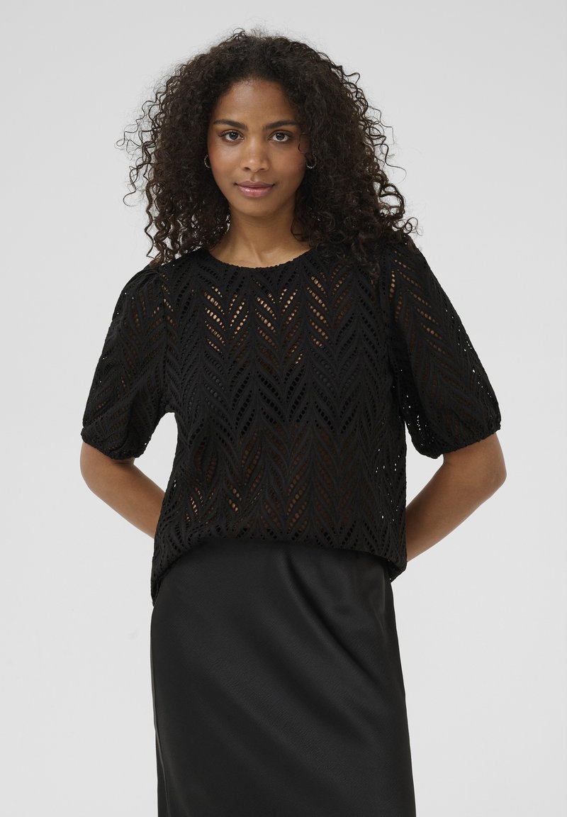 Haut noir à manches courtes avec un motif de feuilles en œillets, présentant un col rond et des manches bouffantes amples. Tissu lisse en dessous.