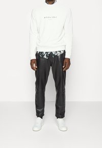 Felpa bianca con il testo "Boglioli Milano", sovrapposta a una camicia a fantasia in bianco e nero, abbinata a pantaloni in pelle neri e sneakers bianche.