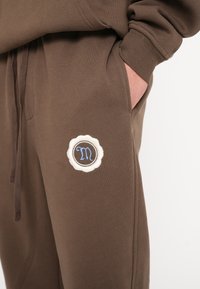 Braune Baumwoll-Jogginghosen mit einer Seitentasche und einem dekorativen Patch mit einem weißen gewellten Rand und dem blauen Buchstaben "M". Glatte Textur.