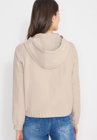 Veste à capuche beige avec poignets et ourlet élastiques, présentant une texture lisse et une coupe décontractée, vue de dos avec un jean en denim.