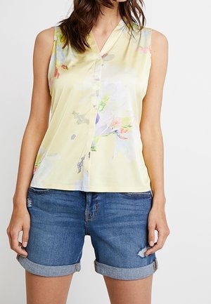 Femme portant un chemisier sans manches jaune clair à fleurs et un short en jean bleu retroussé avec une légère usure sur une jambe.