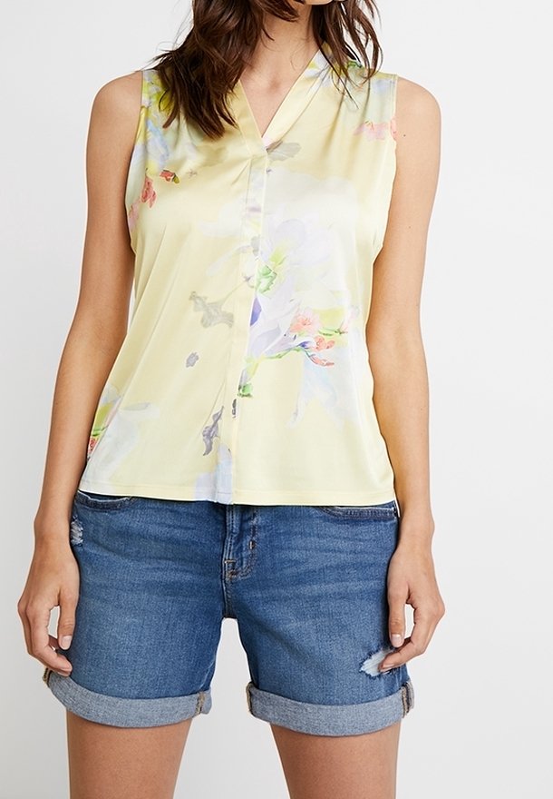 Femme portant un chemisier sans manches jaune clair à fleurs et un short en jean bleu retroussé avec une légère usure sur une jambe.