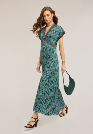 Mujer caminando con un vestido largo de estampado cachemir en tonos verde azulado y blanco, llevando un bolso verde oscuro, sandalias de tacón marrones y joyas doradas.