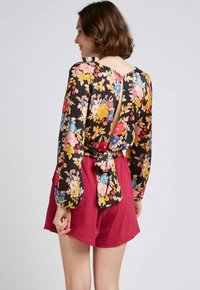 Blouse fleurie à manches longues noire, ornée de fleurs jaunes, roses et bleues ; découpes au dos avec un nœud. Portée avec un short bordeaux.
