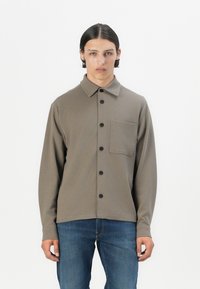Brown long-sleeve button-up shirt with a collar, featuring a chest pocket and black buttons. Smooth texture, simple design, tucked into blue jeans.