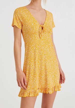Robe jaune à manches courtes avec imprimé floral blanc, détail noué au col en V et ourlet volanté portée par une personne debout.