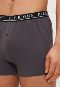 Graue Boxershorts mit glatter Textur, Knopfverschluss und einem schwarzen Bund, auf dem "PIER ONE" in weißer Schrift gedruckt ist.