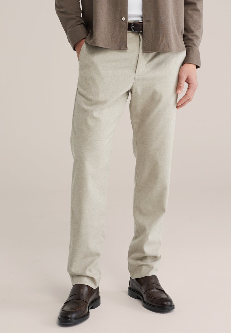 Homme portant un pantalon beige, une ceinture en cuir marron foncé, des mocassins marron et une chemise marron par-dessus un sous-vêtement blanc, debout avec les mains détendues.