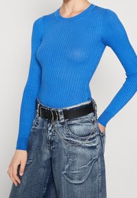 Top blu ribbed a maniche lunghe abbinato a jeans in denim dal fit morbido. Cintura nera con dettagli in catena aggiunge contrasto e funzionalità.