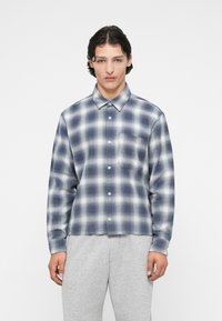 Srajca z mornarsko modro in belo karo s poklopcem na gumbih, dolgimi rokavi ter enim žepom na prsih. Izdelana iz teksturirane tkanine.