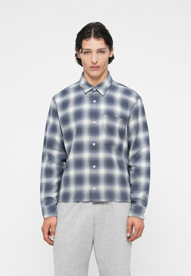 Srajca z mornarsko modro in belo karo s poklopcem na gumbih, dolgimi rokavi ter enim žepom na prsih. Izdelana iz teksturirane tkanine.